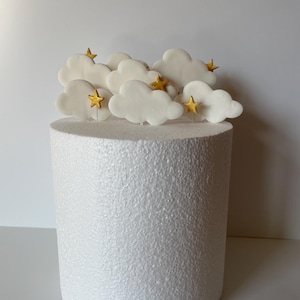 May include: A white cake with cloud-shaped decorations and gold star accents. The cake is cylindrical and sits on a white surface. The cloud and star decorations are arranged on top of the cake.