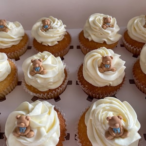 May include: A dozen cupcakes in a white box, each topped with white frosting and a small teddy bear decoration. The cupcakes have a golden-brown base and are arranged in rows.