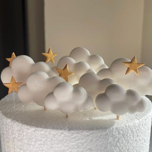 May include: A cake topper featuring white cloud decorations and gold star accents. The clouds are arranged in a cluster, with several gold stars on thin sticks. The cake base is white and textured, creating a whimsical and celebratory design.