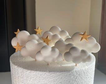 May include: A cake topper featuring white cloud decorations and gold star accents. The clouds are arranged in a cluster, with several gold stars on thin sticks. The cake base is white and textured, creating a whimsical and celebratory design.
