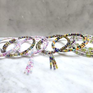 May include: Several beaded coil bracelets in various colors, including pink, purple, green, and blue. Some bracelets have dangling beaded tassels. The bracelets are displayed on a white marble surface.
