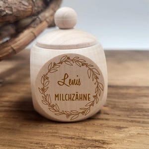 May include: A small, light-colored wooden jar with a lid and a round knob. The jar has a circular sticker with the name "Lenis" and the words "MILCHZ&Auml;HNE" inside a decorative wreath. The jar is sitting on a wooden surface.