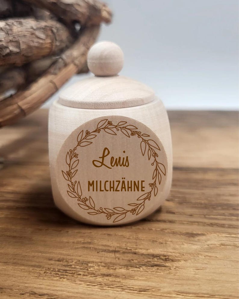 May include: A small, light-colored wooden jar with a lid and a round knob. The jar has a circular sticker with the name "Lenis" and the words "MILCHZ&Auml;HNE" inside a decorative wreath. The jar is sitting on a wooden surface.