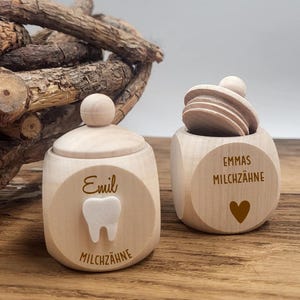 May include: Two wooden tooth keepsake boxes. One box has a lid and the name "Emil" and a tooth graphic. The other box is open and has the name "Emmas" and a heart graphic. The text "Milchzahne" is on both boxes.