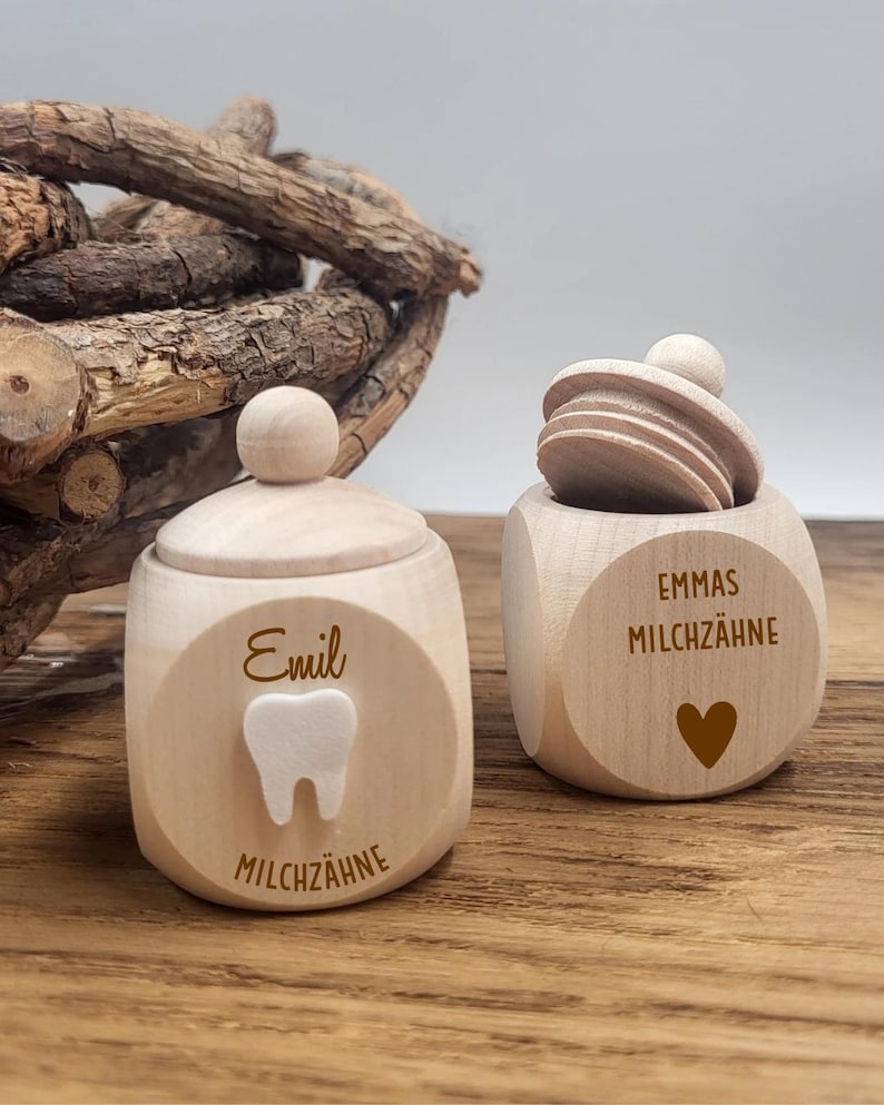 May include: Two wooden tooth keepsake boxes. One box has a lid and the name "Emil" and a tooth graphic. The other box is open and has the name "Emmas" and a heart graphic. The text "Milchzahne" is on both boxes.