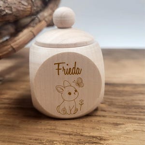 May include: A small, light-colored wooden box with a lid and a round knob. The box features the name "Frieda" and a cute illustration of a bunny and butterfly. The box is sitting on a wooden surface.