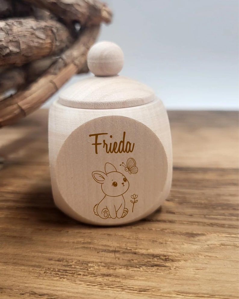 May include: A small, light-colored wooden box with a lid and a round knob. The box features the name "Frieda" and a cute illustration of a bunny and butterfly. The box is sitting on a wooden surface.