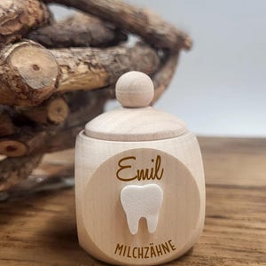 May include: A small, light-colored wooden tooth fairy box with a lid and a round knob. The box has a circular emblem with the name "Emil", a tooth graphic, and the word "MILCHZ&Auml;HNE".