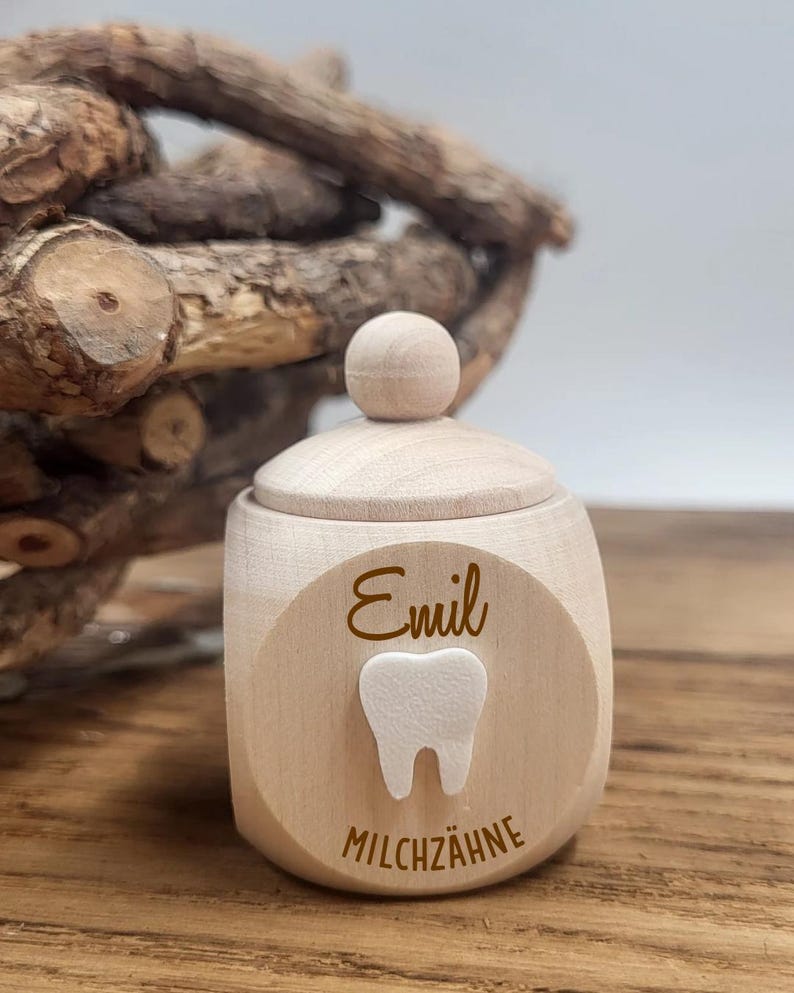 May include: A small, light-colored wooden tooth fairy box with a lid and a round knob. The box has a circular emblem with the name "Emil", a tooth graphic, and the word "MILCHZ&Auml;HNE".