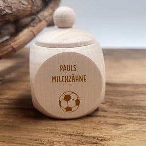May include: A small, light-colored wooden box with a lid and a spherical knob. The box features the text "PAULS MILCHZ&Auml;HNE" and a soccer ball graphic on the front. The box is sitting on a wooden surface.