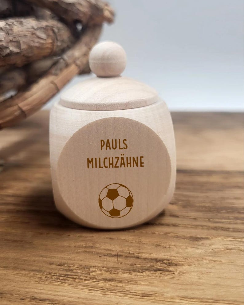 May include: A small, light-colored wooden box with a lid and a spherical knob. The box features the text "PAULS MILCHZ&Auml;HNE" and a soccer ball graphic on the front. The box is sitting on a wooden surface.