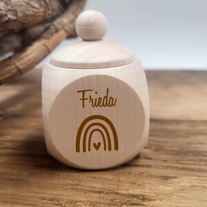 May include: A small, light-colored wooden box with a lid and a round knob. The box has the name "Frieda" and a rainbow design printed on the front. The box is sitting on a wooden surface.