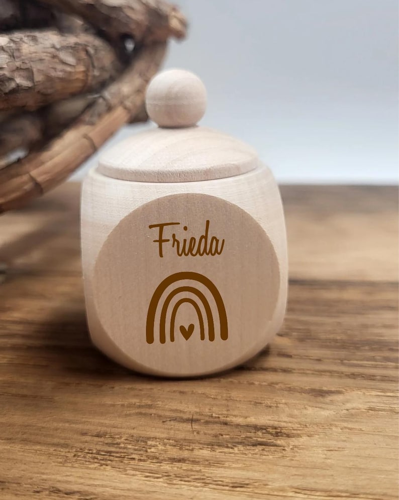 May include: A small, light-colored wooden box with a lid and a round knob. The box has the name "Frieda" and a rainbow design printed on the front. The box is sitting on a wooden surface.