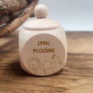 May include: A small, light-colored wooden box with a lid and a round knob on top. The box has a circular label with the text "EMMAS MILCHZ&Auml;HNE" and illustrations of a lion, elephant, and giraffe. The box is sitting on a wooden surface.