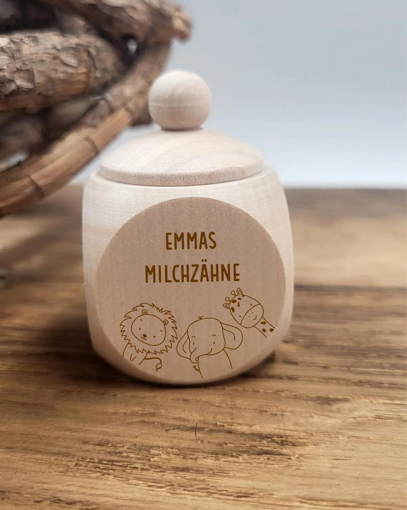 May include: A small, light-colored wooden box with a lid and a round knob on top. The box has a circular label with the text "EMMAS MILCHZ&Auml;HNE" and illustrations of a lion, elephant, and giraffe. The box is sitting on a wooden surface.