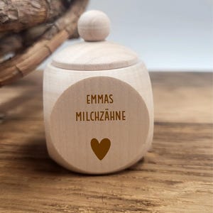 May include: A small, light-colored wooden box with a lid and a round knob on top. The box has a circular label with the text "EMMAS MILCHZ&Auml;HNE" and a heart symbol. The box is sitting on a wooden surface.