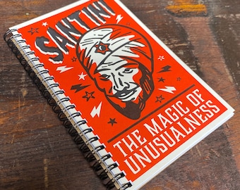 May include: A spiral-bound notebook with a red cover featuring the name "SANTINI" in large white letters. The cover also displays an illustration of a man in a turban and the text "THE MAGIC OF UNUSUALNESS".
