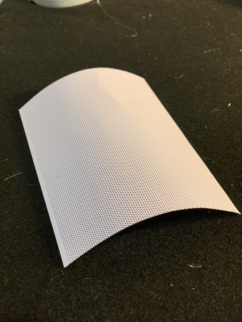 May include: A curved, white perforated sheet of material, possibly fabric or paper, with a grid of small holes. The sheet is positioned on a dark, textured surface, creating a contrast between the light and dark tones.