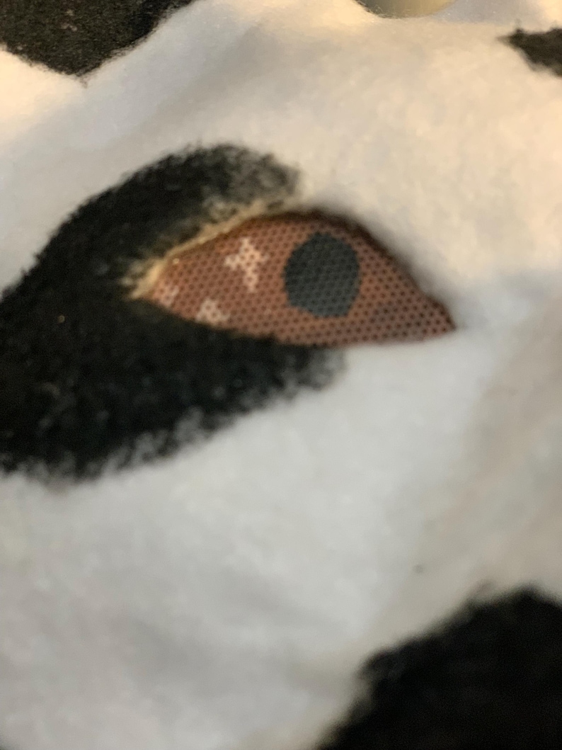 May include: Close-up of a plush toy eye. The eye is a light brown color with a black pupil. The eye is surrounded by black fur and set against a white background.