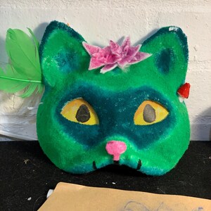 May include: A green cat mask with a pink nose and a pink flower on top. The mask has yellow mesh eyes and a green feather on the side. The mask is made of a soft, fuzzy material.