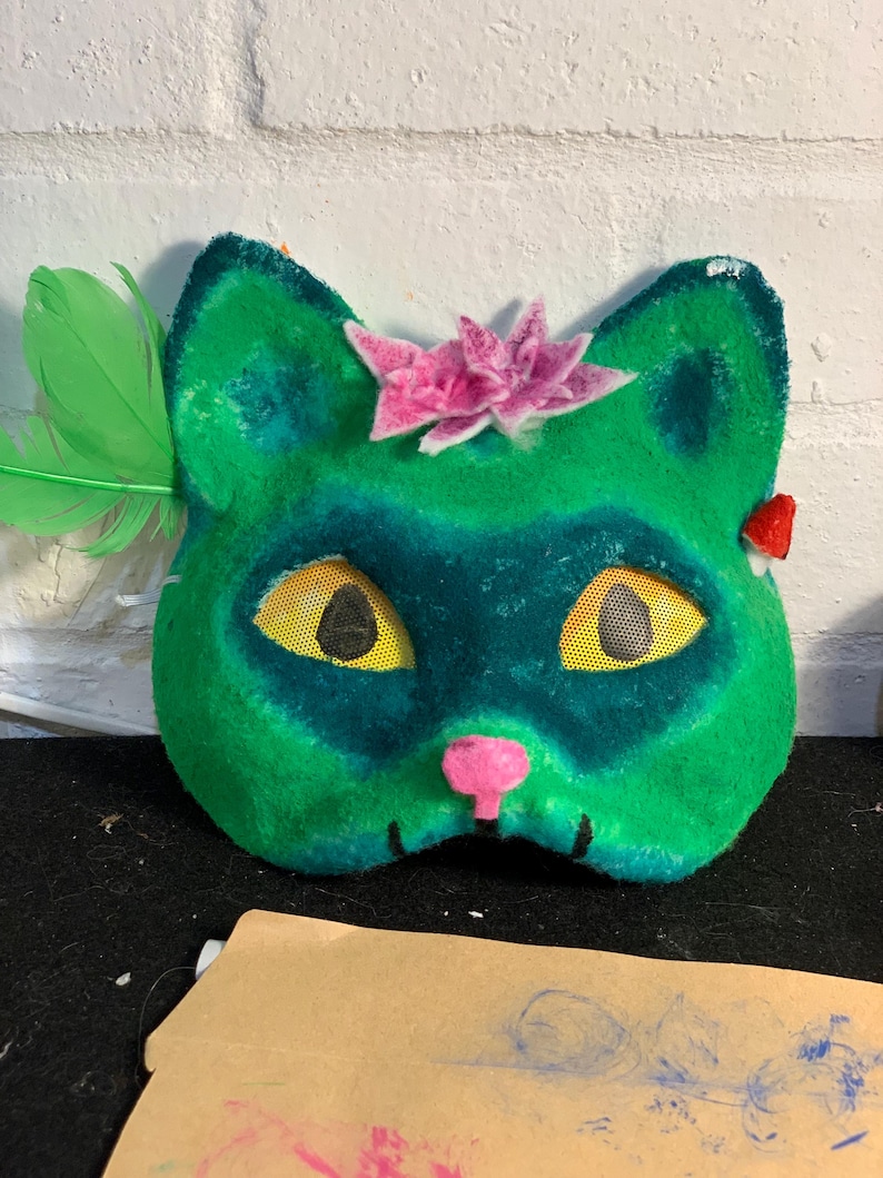 May include: A green cat mask with a pink nose and a pink flower on top. The mask has yellow mesh eyes and a green feather on the side. The mask is made of a soft, fuzzy material.