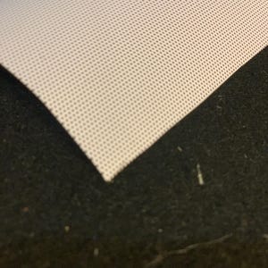 May include: A close-up of a white, perforated fabric with a grid of small holes. The fabric is folded over, revealing a dark, textured surface beneath. The fabric's edge is visible, showing its thickness and texture.