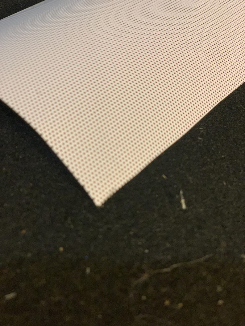 May include: A close-up of a white, perforated fabric with a grid of small holes. The fabric is folded over, revealing a dark, textured surface beneath. The fabric's edge is visible, showing its thickness and texture.