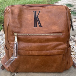 May include: A brown faux leather backpack with a gold zipper and a black embroidered letter "K" on the front.