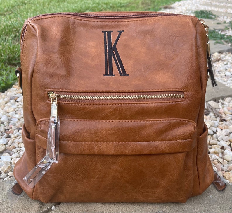 May include: A brown faux leather backpack with a gold zipper and a black embroidered letter "K" on the front.