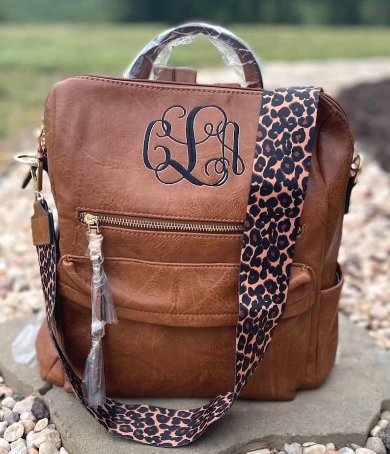 May include: A brown faux leather backpack with a monogrammed front pocket and a leopard print adjustable strap.