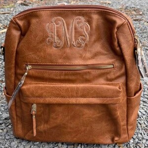 May include: A brown faux leather backpack with a gold zipper and a monogrammed "MCS" on the front.