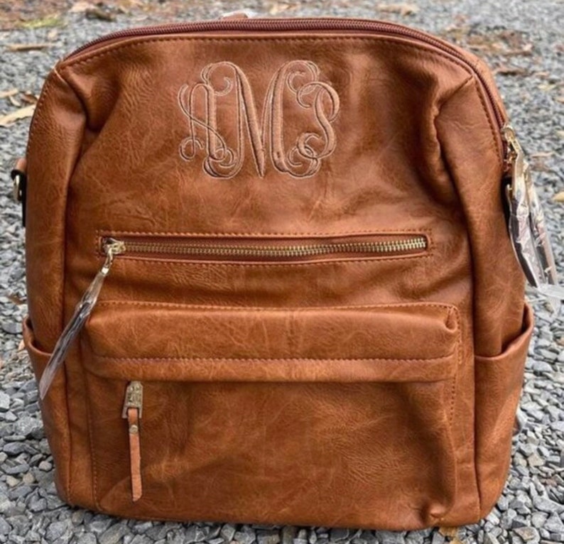 May include: A brown faux leather backpack with a gold zipper and a monogrammed "MCS" on the front.