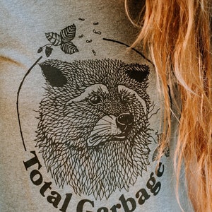 K&ouml;nnte beinhalten: Ein graues T-Shirt mit einer schwarz-wei&szlig;en Illustration eines Tierkopfes. Die Illustration ist von einem Kreis umgeben, auf dem der Text "Total Garbage" steht.