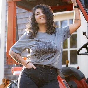 K&ouml;nnte beinhalten: Eine Frau tr&auml;gt ein graues T-Shirt mit einem schwarz-wei&szlig;en Waschb&auml;r-Motiv und dem Text "Total Garbage". Sie steht neben einem roten Traktor.