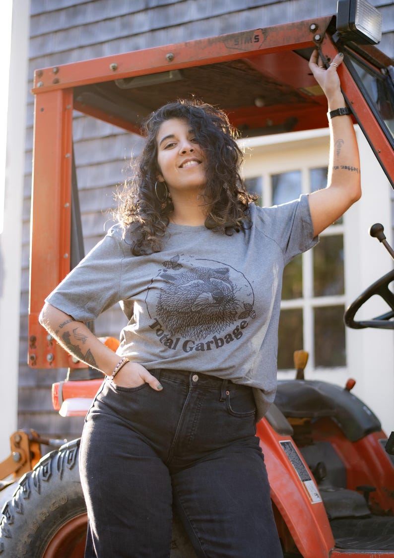 K&ouml;nnte beinhalten: Eine Frau tr&auml;gt ein graues T-Shirt mit einem schwarz-wei&szlig;en Waschb&auml;r-Motiv und dem Text "Total Garbage". Sie steht neben einem roten Traktor.