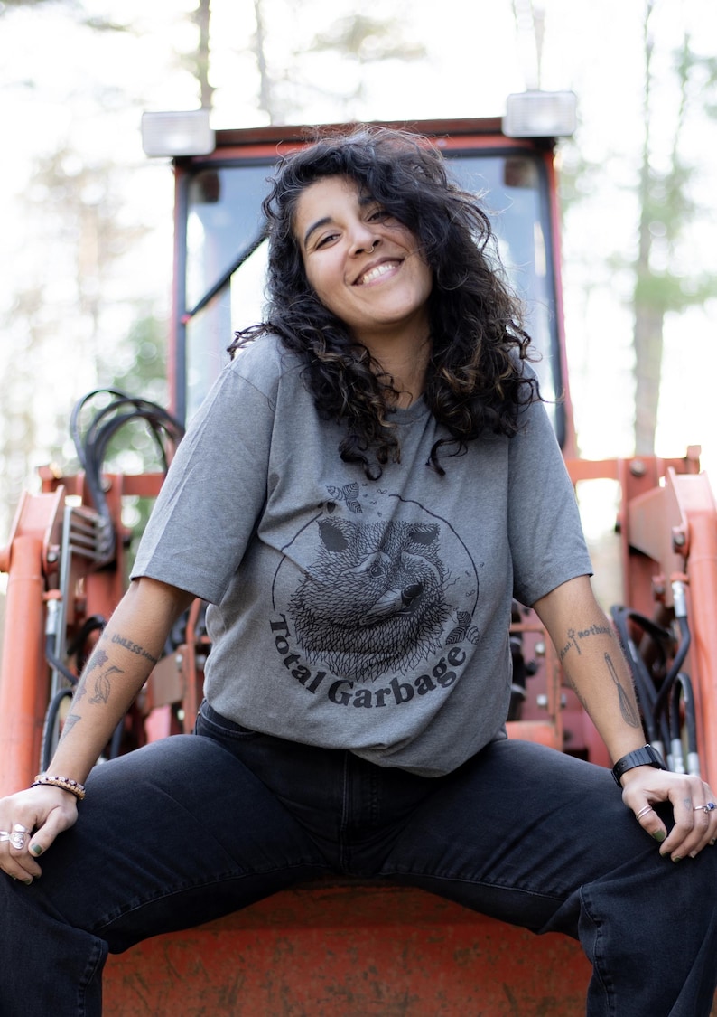 K&ouml;nnte beinhalten: Eine Person tr&auml;gt ein graues T-Shirt mit einem schwarz-wei&szlig;en Waschb&auml;ren-Motiv und dem Text "Total Garbage". Die Person sitzt auf einem roten Ger&auml;t.