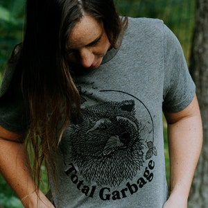 K&ouml;nnte beinhalten: Ein heidefarbenes T-Shirt mit einer schwarz-wei&szlig;en Illustration eines Waschb&auml;renkopfes. Der Text "Total Garbage" ist in Schwarz unter der Illustration aufgedruckt.