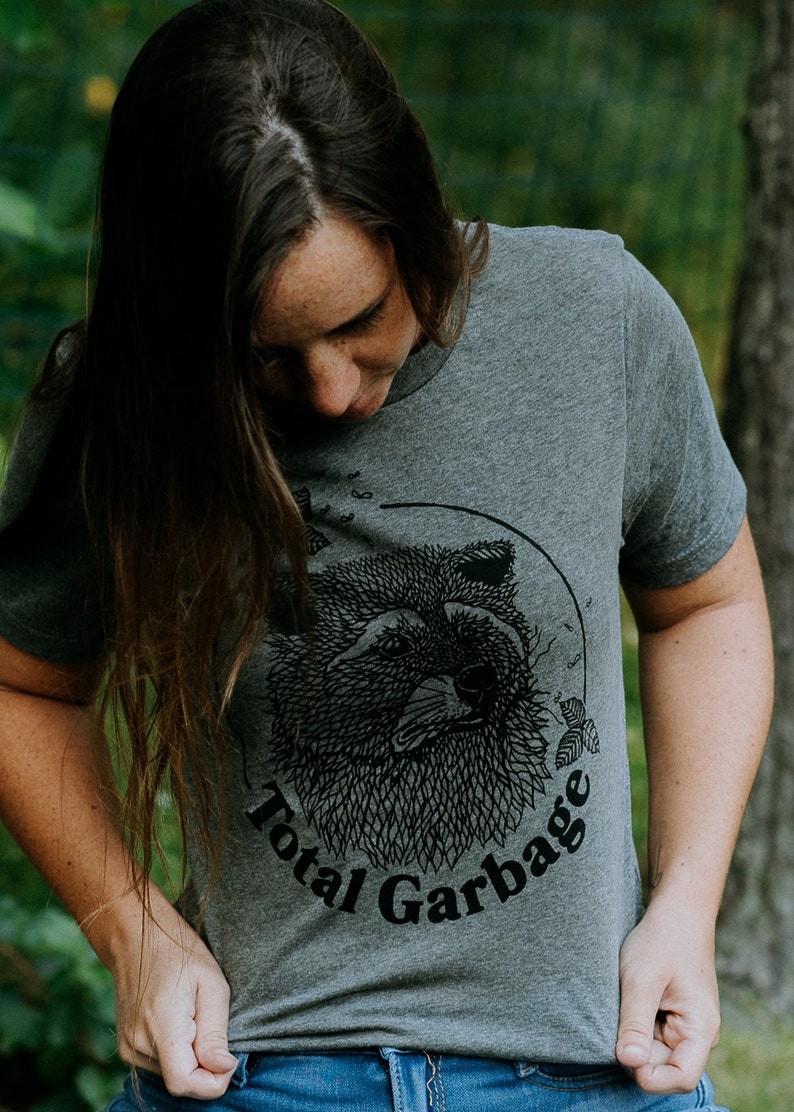 K&ouml;nnte beinhalten: Ein heidefarbenes T-Shirt mit einer schwarz-wei&szlig;en Illustration eines Waschb&auml;renkopfes. Der Text "Total Garbage" ist in Schwarz unter der Illustration aufgedruckt.