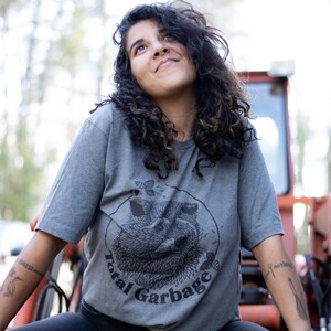 K&ouml;nnte beinhalten: Eine Person tr&auml;gt ein graues T-Shirt mit einem schwarz-wei&szlig;en Bild eines Waschb&auml;ren in einem Kreis mit dem Text "Total Garbage".