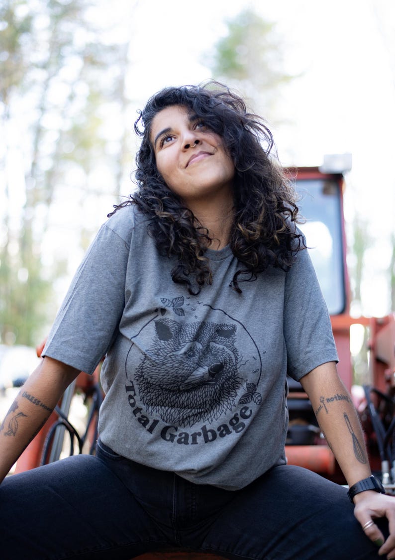 K&ouml;nnte beinhalten: Eine Person tr&auml;gt ein graues T-Shirt mit einem schwarz-wei&szlig;en Bild eines Waschb&auml;ren in einem Kreis mit dem Text "Total Garbage".