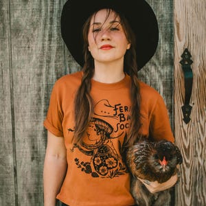 May include: A person wearing a brown t-shirt with a black and white illustration of a woman wearing a hat and holding a flower. The shirt has the text "Ferndale Blacksmith Society" printed on it. The person is holding a brown and black chicken.