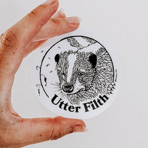 May include: A white circular sticker with a black and white illustration of a badger's face. The text "Utter Filth" is printed below the badger's face.