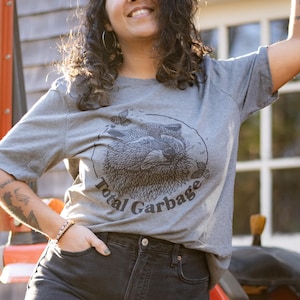 K&ouml;nnte beinhalten: Eine Frau tr&auml;gt ein graues T-Shirt mit einem Waschb&auml;r-Motiv und dem Text "Total Garbage". Sie tr&auml;gt eine schwarze Jeans und hat lockiges braunes Haar.
