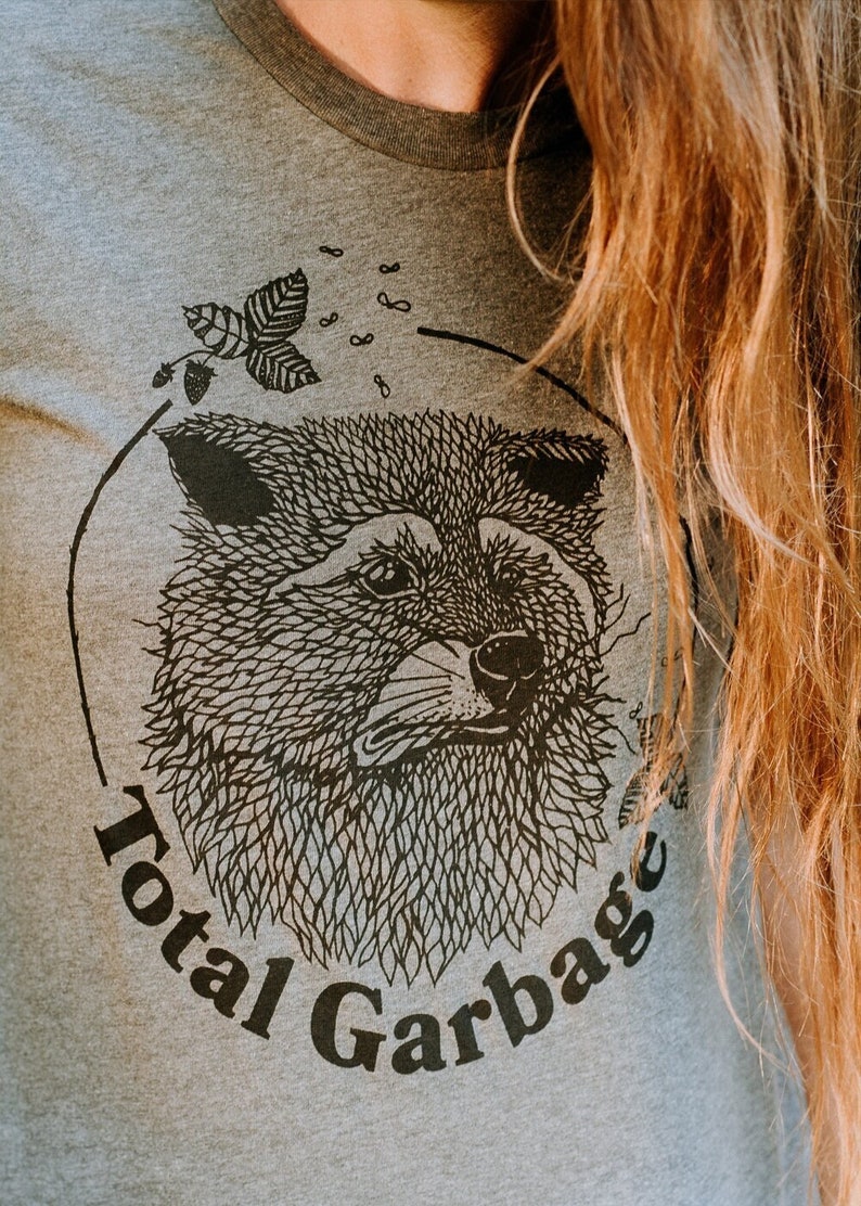 K&ouml;nnte beinhalten: Ein graues T-Shirt mit einer schwarz-wei&szlig;en Illustration eines Tierkopfes. Die Illustration ist von einem Kreis umgeben, auf dem der Text "Total Garbage" steht.