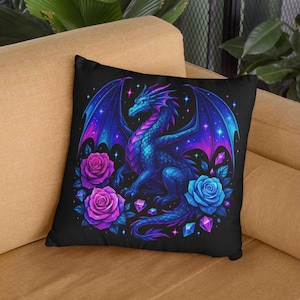 May include: A square throw pillow featuring a black background with a central illustration of a blue and purple dragon with large wings. The dragon is surrounded by vibrant pink and blue roses, scattered geometric crystals, and small glowing stars.