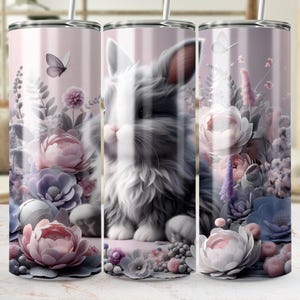 May include: A stainless steel tumbler featuring a design of a fluffy gray and white rabbit surrounded by pastel-colored flowers and butterflies. The floral arrangement includes pink, purple, and blue blooms. The tumbler has a straw and a light pink background.