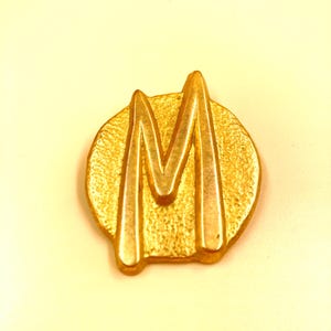 May include: A gold-coloured metal pin featuring the letter "M" in a stylized design. The letter is raised and has a textured surface, set against a textured circular background. The pin has a shiny, metallic finish.