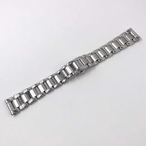 May include: A silver-toned metal watch band featuring a series of polished rectangular links connected in a classic three-piece design. The strap includes a secure folding clasp mechanism and a smooth, reflective finish throughout the entire length.