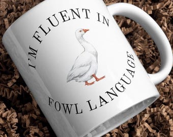 May include: White ceramic mug with a goose illustration and the text "I'M FLUENT IN FOWL LANGUAGE". The mug is inside a white box filled with brown crinkle paper.