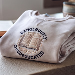 Könnte beinhalten: Ein gefalteter beigefarbener Sweatshirt mit dem gestickten Text "DANGEROUSLY OVEREDUCATED" um ein offenes Buchdesign. Der Sweatshirt ist ordentlich auf einer hellen Oberfläche gefaltet, mit einem Buch und einer Tasse im Hintergrund.